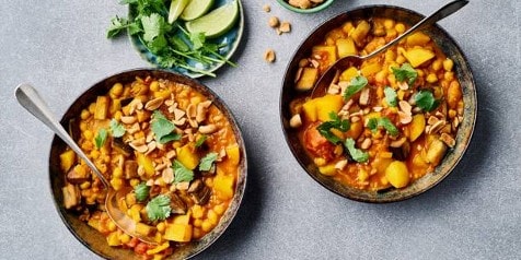 Twee kommen met curry van kikkererwten en groenten, gegarneerd met koriander en pinda's, naast een bordje met limoenpartjes en koriander.