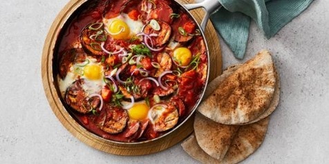Shakshuka in een pan met ongekookte eieren en gegrilde groenten, geserveerd met pita broodjes.
