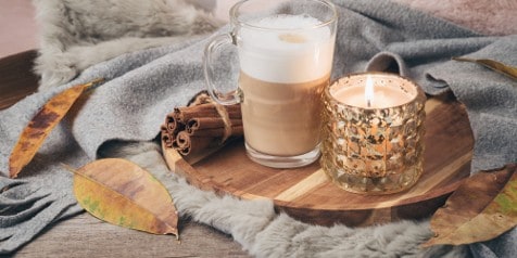 Herfstachtige setting met een kopje cappuccino, kaneelstokjes en een brandende kaars op een houten dienblad, omringd door decoratieve bladeren en zachte dekens.
