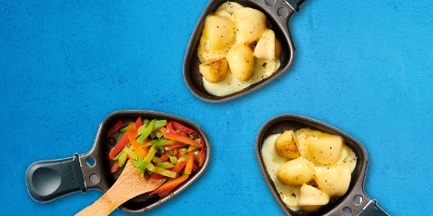 Raclette pannetjes met gesmolten kaas en aardappelen, en een houten lepeltje met gesneden paprika op een blauwe achtergrond.