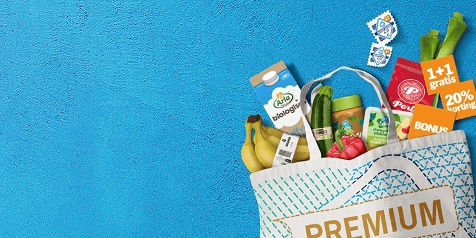 Boodschappentas met diverse producten waaronder Arla biologische melk, bananen, komkommer, paprika, Perla koffie, en bonus stickers met 1+1 gratis en 20% korting op een blauwe achtergrond.
