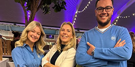 Drie lachende Albert Heijn medewerkers in blauwe werkkleding, staand met gekruiste armen onder decoratieve verlichting in de winkel.