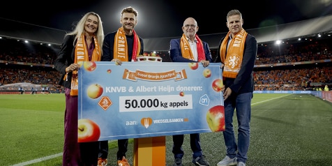 Grote cheque met de tekst 'KNVB & Albert Heijn doneren: 50.000 kg appels aan Voedselbanken Nederland', vastgehouden door vier mensen in voetbalstadion, decoratief met oranje sjaals en Nederlandse vlagkleuren, uitreiking tijdens evenement.