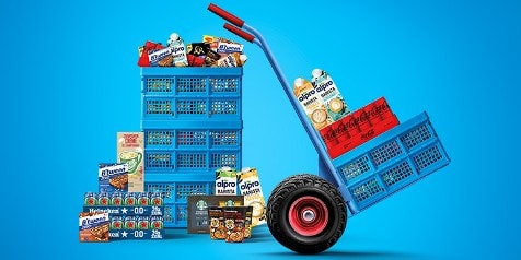Blauwe steekwagen en kratten gevuld met producten, waaronder Coca-Cola, Heineken, Alpro, B’tween en Lassie rijst, tegen een blauwe achtergrond.