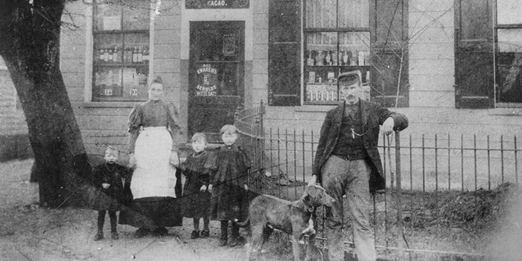 Oude zwart-wit foto van een gezin met drie kinderen en een hond voor een winkelingang.