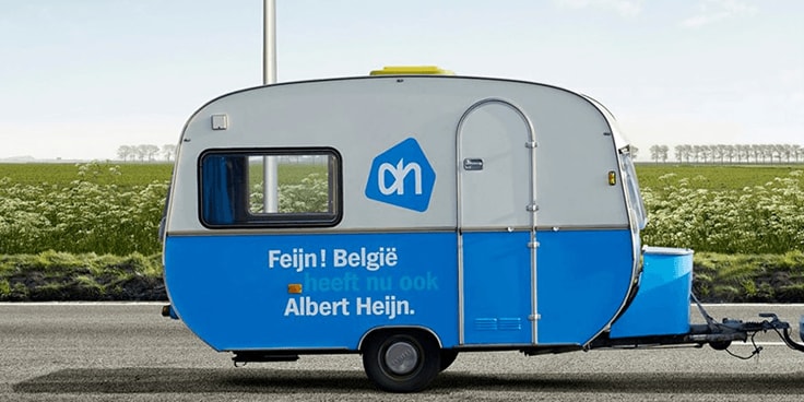 Caravan met Albert Heijn-logo en tekst "Feijn! België heeft nu ook Albert Heijn." langs een weg met graslandschap op de achtergrond.