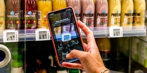 Hand die een smartphone gebruikt om een product te scannen in een Albert Heijn winkel, met smoothies en proteïne drankjes op de achtergrond.