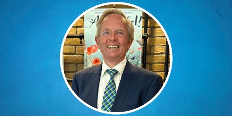 Man in pak met blauwe achtergrond en bloemenposter op de achtergrond.