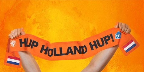 Twee handen houden een oranje sjaal omhoog met de tekst "Hup Holland Hup!", het Albert Heijn-logo en Nederlandse vlaggetjes, tegen een oranje achtergrond.