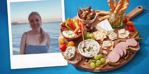 Borrelschotel met kaas, worst, crackers, druiven, dips en groentesticks op een houten snijplank naast een wazige foto van een vrouw met een zee als achtergrond.