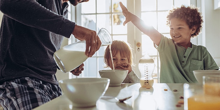 Ontbijtscène met vader die melk uitschenkt in een kom, zittend aan tafel met twee kinderen.
