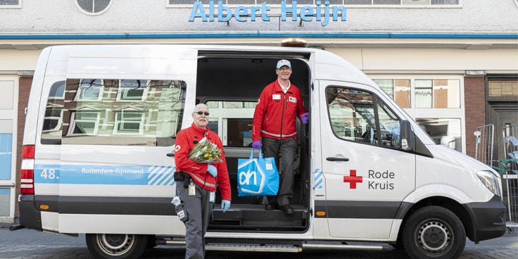 Albert Heijn busje voor een filiaal, met Rode Kruis medewerkers die boodschappen van Albert Heijn en een bos bloemen uitladen.