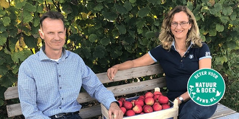 Twee mensen zitten op een bankje naast een houten kist met rode appels, met een "Beter voor Natuur & Boer" keurmerk zichtbaar. Op de achtergrond zijn groene planten.