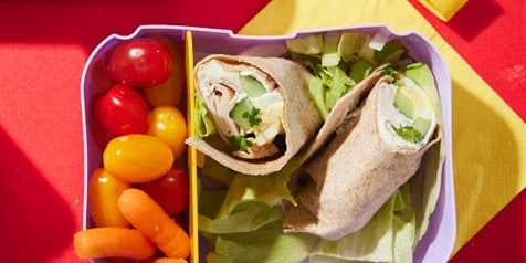 Twee volkoren wraps gevuld met kip, sla en komkommer in een lunchbox, geserveerd met snoeptomaatjes en babywortels.