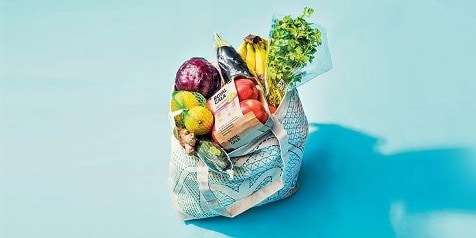 Herbruikbare boodschappentas gevuld met diverse groenten en fruit op een blauwe achtergrond.