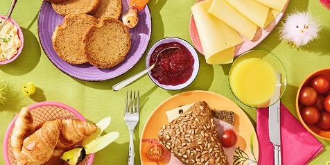 Ontbijttafel met volkorenbrood, croissant, schijfjes kaas, jam, sinaasappelsap en kerstomaatjes.