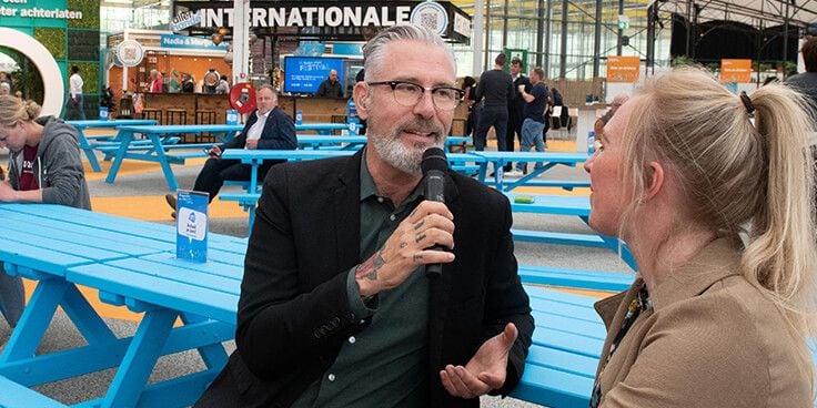Mensen in gesprek op een evenement met blauwe picknicktafels op de achtergrond.