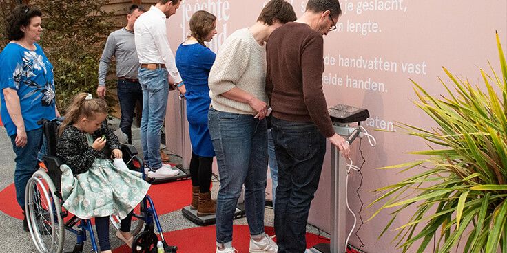 Mensen in de rij voor een activiteit, waaronder een kind in een rolstoel, naast een roze achtergrond met tekst.