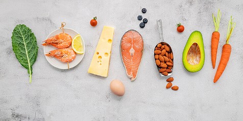 Diverse selectie voedingsmiddelen, waaronder boerenkool, garnalen, kaas, zalmfilet, amandelen, avocado, wortelen, een ei, bosbessen en cherrytomaten.