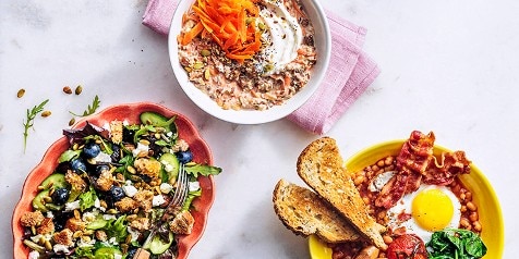 Drie ontbijtborden met diverse gerechten: een salade met bosbessen en geitenkaas, een kommetje havermout met geraspte wortel en zaden en een bord met spek, gebakken ei, bonen en geroosterd brood.