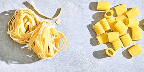 Twee soorten ongekookte pasta op een stenen ondergrond, links lintpasta en rechts korte pastabuisjes.