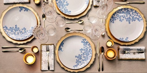 Gedekte tafel met wit porselein met blauwe bloemen en gouden rand, kristallen glazen, gouden bestek en kaarsen.