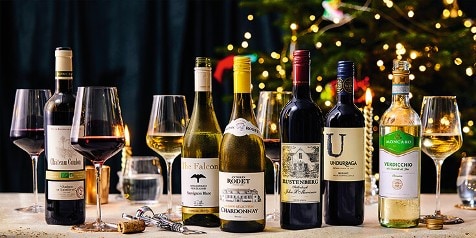 Assortiment van wijnflessen en gevulde wijnglazen op een gedecoreerde tafel met kerstverlichting op de achtergrond.