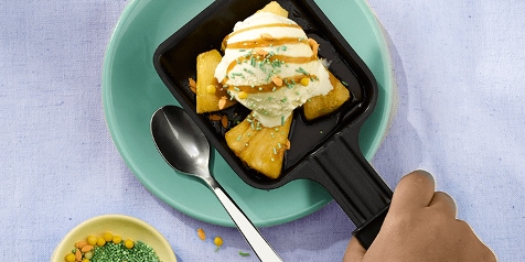 Ananasdessert met vanille-ijs en karamelsaus in een zwarte schaal, geserveerd op een groen bord, met een hand die de schaal vasthoudt en een lepel ernaast.