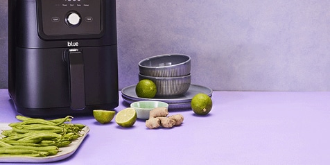 Airfryer met verse ingrediënten zoals sperziebonen, gember, en limoenen op een aanrecht.