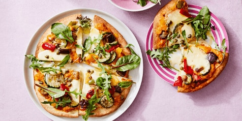 Pizza met groenten en rucola op borden, geserveerd op een lichtpaarse achtergrond.