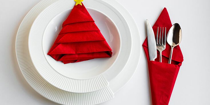 Decoratief gevouwen servet in de vorm van een kerstboom op een wit bord met bestek in een rode servetwikkel.