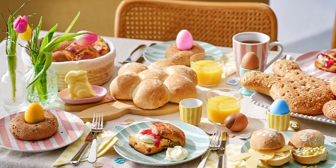Paastafel met diverse broodjes, gekleurde eieren, een konijntje van boter, sinaasappelsap en tulpen in een vaas.
