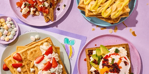 Verschillende wafels met diverse toppings waaronder aardbeien met room, avocado met ei en tomaat met feta, geserveerd op gekleurde borden.