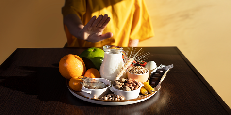 Person voor een tafel met diverse voedingsmiddelen zoals melk, noten, eieren en vruchten, waarbij een handgebaar van afwijzing wordt gemaakt.