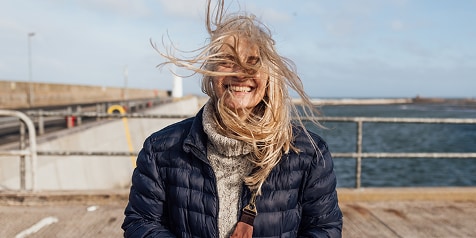 Person met blond haar en een blauwe jas, staande op een pier met de zee op de achtergrond.