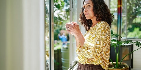 Vrouw in een geel-wit patroon shirt kijkt uit het raam en houdt een beker vast, met groene tuin op de achtergrond