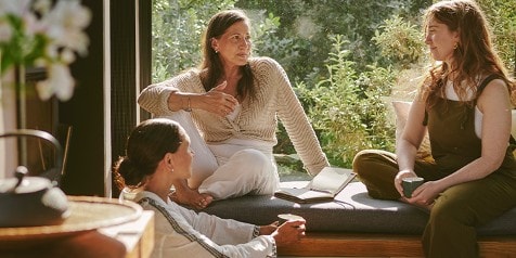 Drie mensen zitten samen op een zonnige vensterbank en voeren een gesprek, met notitieboekjes en een kopje bij de hand