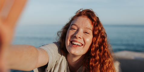 Persoon met rood krullend haar bij zee in selfie pose