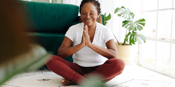Persoon in meditatiehouding met gekruiste benen, zittend op een vloerkleed, met groene planten en een bank op de achtergrond.