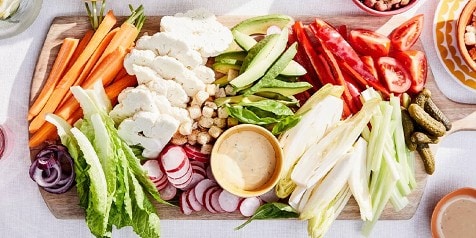 Verse groenteschotel met wortels, bloemkool, avocado, paprika, tomaten, selderij, en dip op een houten plank.