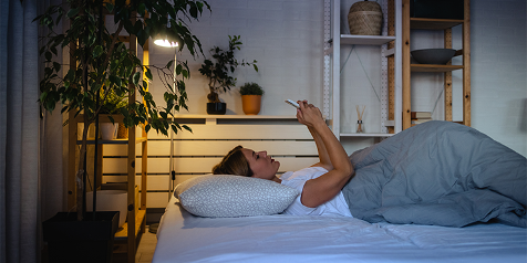 Vrouw liggend in bed terwijl ze op haar telefoon kijkt, omgeven door kamerplanten en sfeerverlichting.
