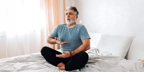 Man in casual kleding mediteert zittend op bed in een lichte kamer.