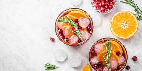 Twee glazen met verfrissende drankjes, gegarneerd met schijfjes sinaasappel, rozemarijn en granaatappelpitten, met een halve sinaasappel en een schaaltje granaatappelpitten op de achtergrond.