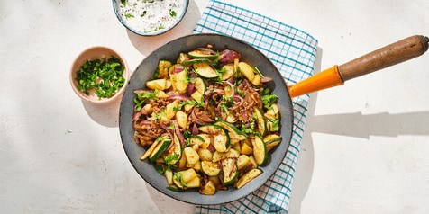 Gebakken aardappelen met groenten en kruiden in een pan, geserveerd met sauzen en verse kruiden op een keukenhanddoek.