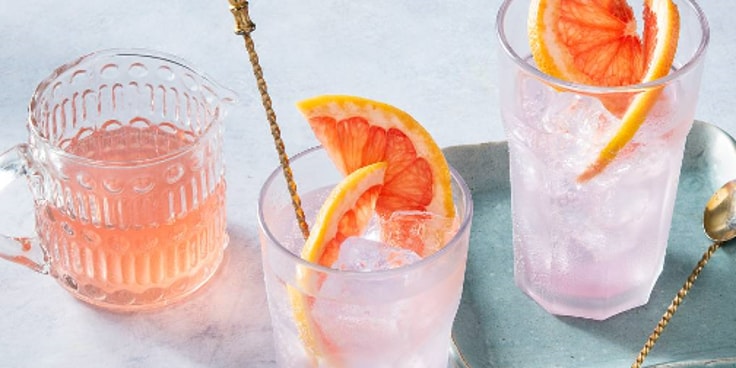 Twee glazen roze grapefruitlimonade met ijsklontjes en schijfjes grapefruit, naast een glazen karaf.