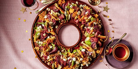 Feestelijke charcuteriekrans met diverse vleeswaren, groenten en kazen op een houten plateau, gedecoreerd met gouden sterretjes.