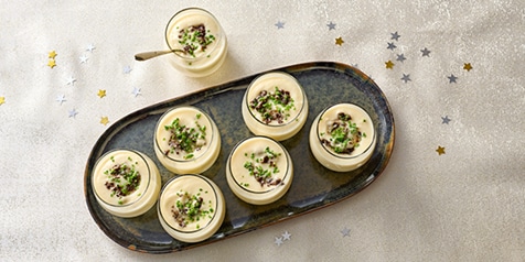 Zeven glazen dessertbekers gevuld met een romig dessert, gegarneerd met groene kruiden, op een donkere schaal geplaatst op een feestelijke tafel met stervormige confetti.