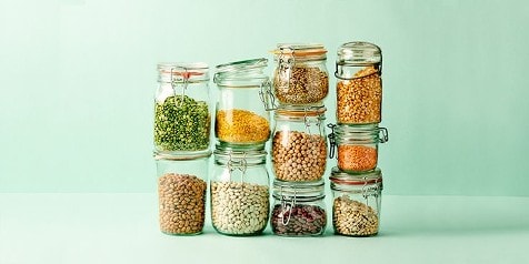 Een stapel glazen potten gevuld met diverse peulvruchten en granen op een mintgroene achtergrond.