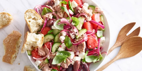 Salade met tonijn, watermeloen, komkommer, rode ui, feta, olijven en verse basilicum geserveerd met knapperig brood op een marmeren tafel.