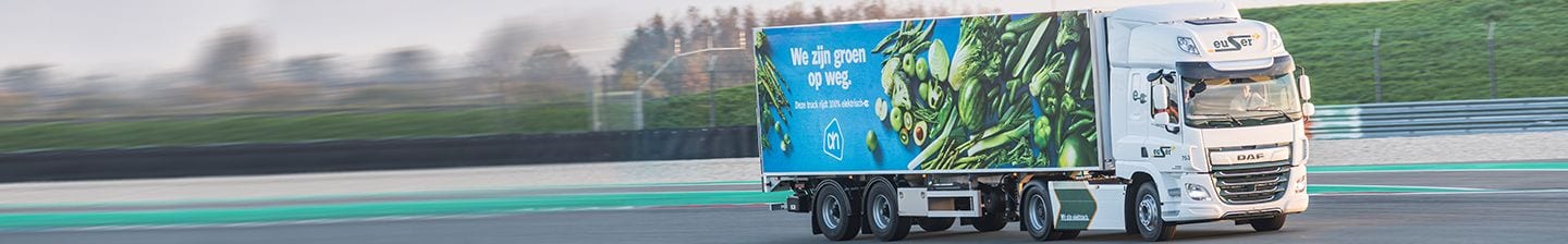 Albert Heijn vrachtwagen met groene groente opdruk en tekst 'We zijn groen op weg' rijdend op de weg.